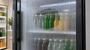 Condensation droplets on the outside of a commercial refrigerator glass door, with a clear wiped patch revealing bottles inside.